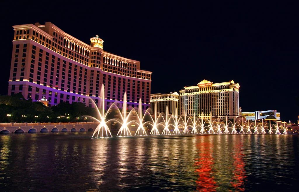 Illuminated Bellagio Fountains performing at night in front of the Bellagio hotel, with nearby Caesars Palace towers in the background.