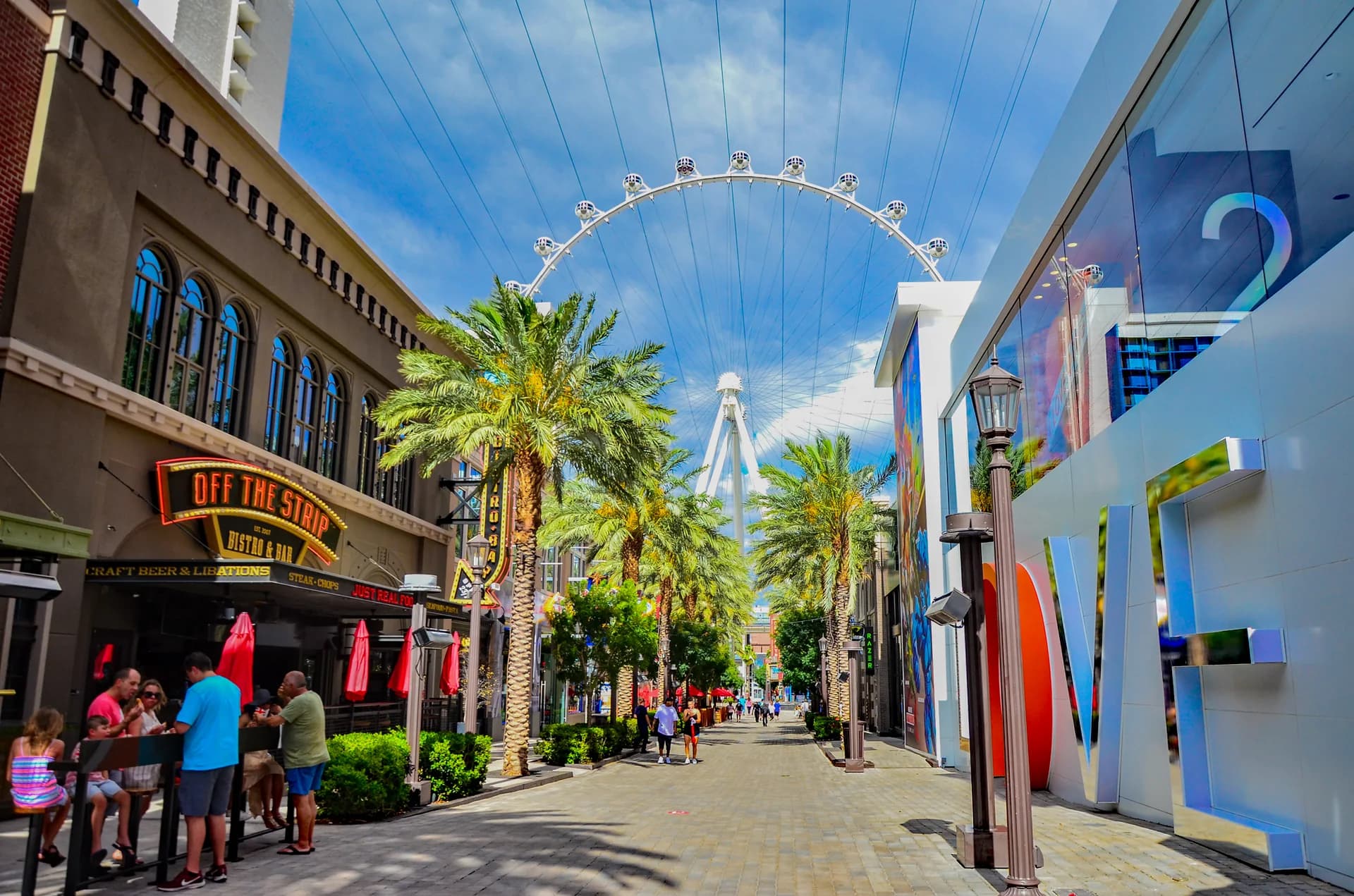 Palm-lined LINQ Promenade pedestrian walkway in daylight, with restaurant seating and the High Roller observation wheel rising behind it.