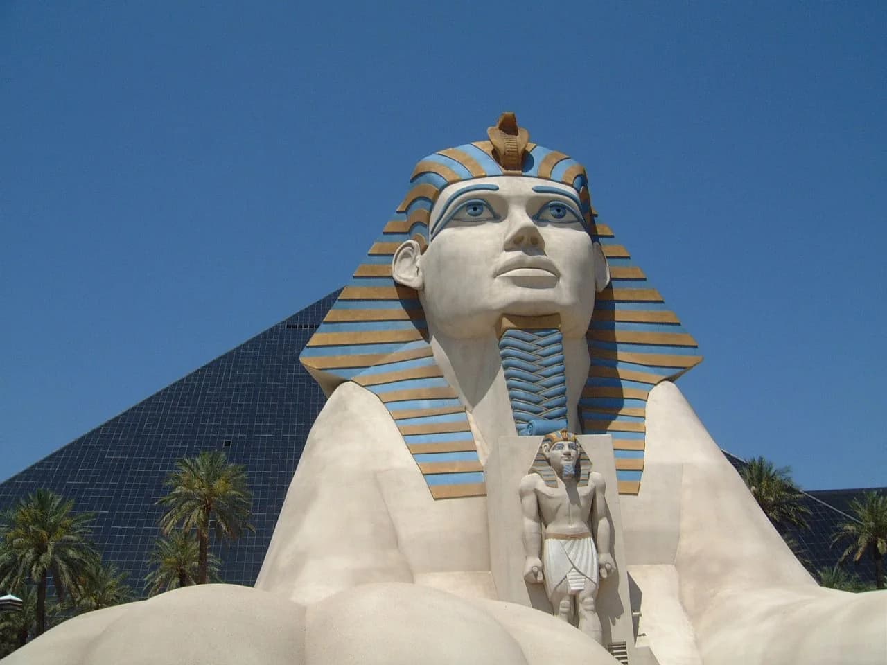 Daytime frontal view of Luxor’s cream-and-blue Sphinx replica with the dark glass pyramid behind it.