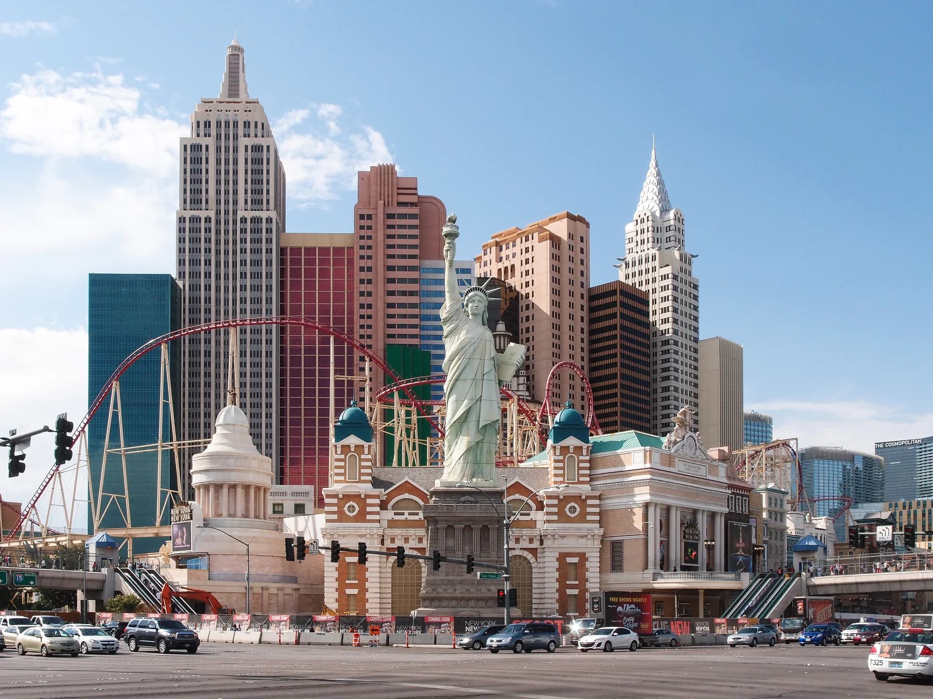 Daytime wide exterior view of New York-New York in Las Vegas, showing the Statue of Liberty replica, skyscraper-style hotel towers, and the Big Apple Coaster.