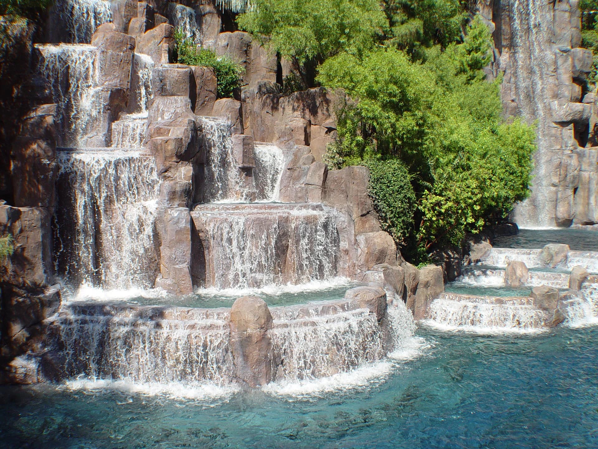 Daytime view of the Lake of Dreams water feature and artificial waterfall at Wynn Las Vegas, with trees and landscaped rock around the water.